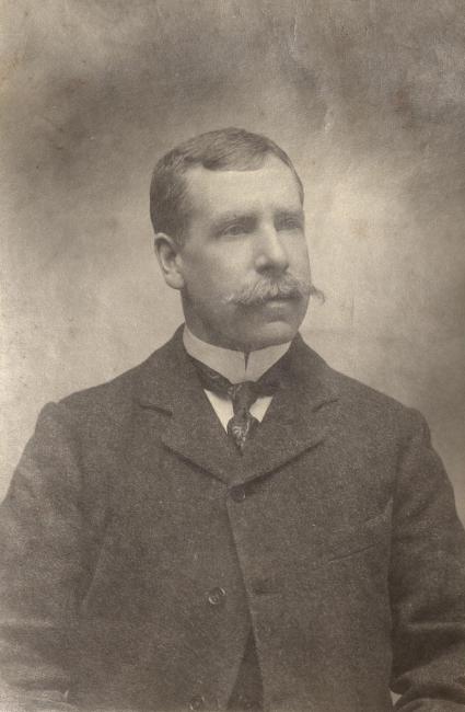 Monochrome photograph of Aloysius O'Kelly. The three quarter length portrait documents him in a jacket, white shirt and mustache. 